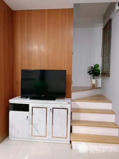 Elegant living space with warm wooden paneling and modern TV cabinet, complemented by sleek marble-like finishes and a cozy staircase leading to an upper level.