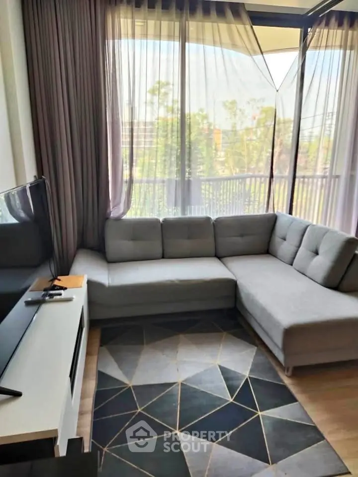 Modern living room with stylish gray sectional sofa and geometric rug, featuring large windows with sheer curtains.