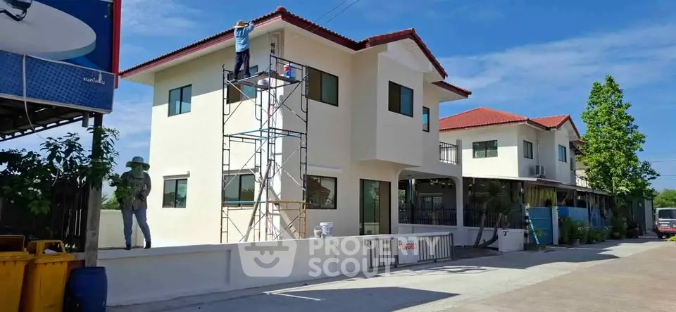 Modern two-story house with red roof under construction, featuring scaffolding and workers, in a sunny neighborhood.