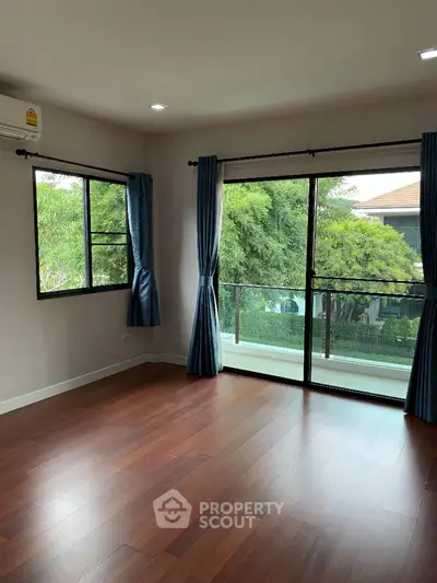 Spacious empty room with wooden flooring and large windows overlooking greenery.