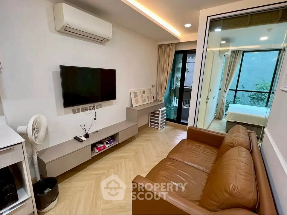 Modern living room with leather sofa and wall-mounted TV, leading to a bedroom with balcony view.