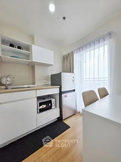 Modern kitchen with sleek white cabinets, compact fridge, and dining area in bright apartment.