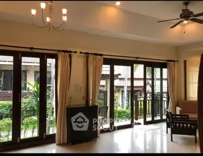 Spacious living room with large windows and garden view, featuring elegant lighting and ceiling fan.