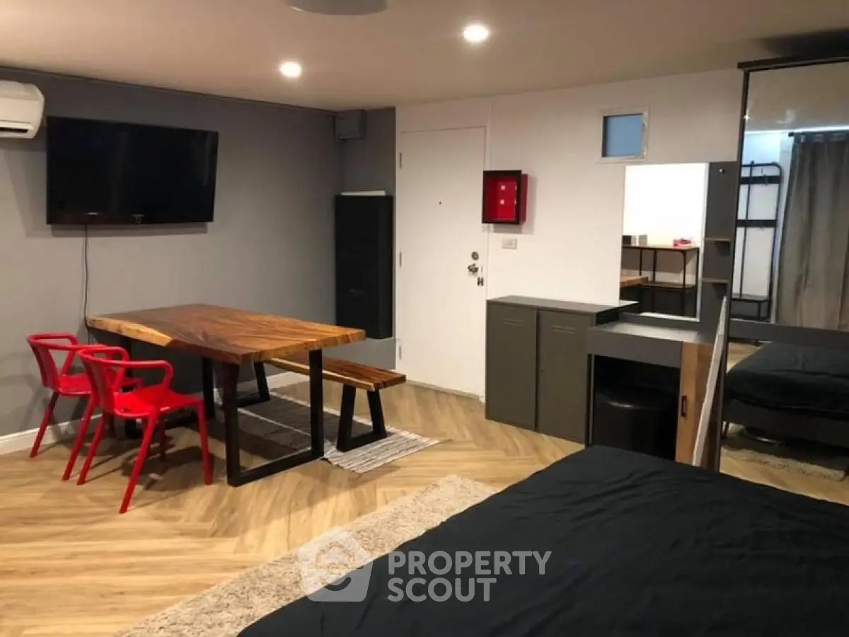 Modern studio apartment with stylish wooden dining table and red chairs, featuring a cozy bed and wall-mounted TV.