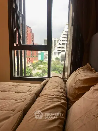 Cozy bedroom with city view through large window, featuring modern bedding and urban skyline.