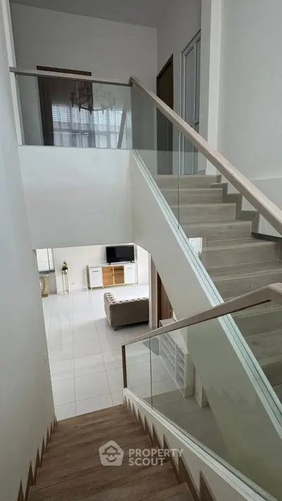 Modern staircase with glass railing leading to a bright living room with sleek furniture.