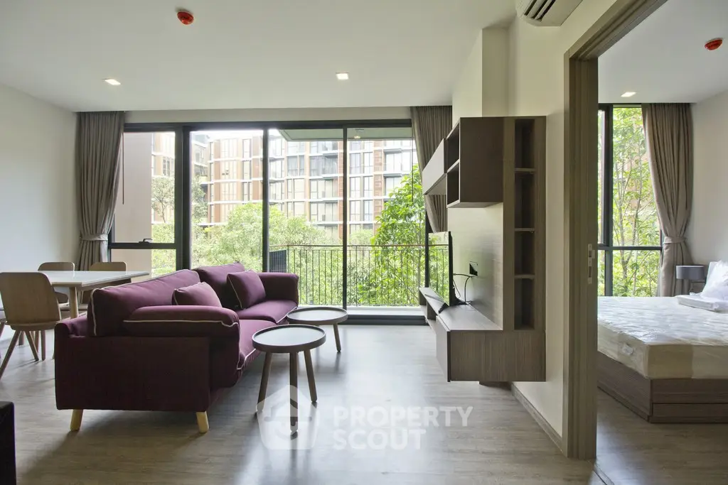 Modern living room with open layout and large windows offering a lush green view, adjacent to a cozy bedroom.