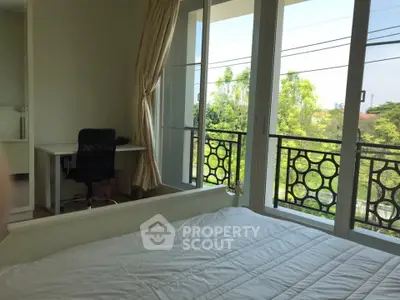 Bright bedroom with large windows and balcony overlooking lush greenery, featuring a cozy workspace.