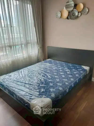 Modern bedroom with large window and stylish decor, featuring a new mattress and wooden flooring.