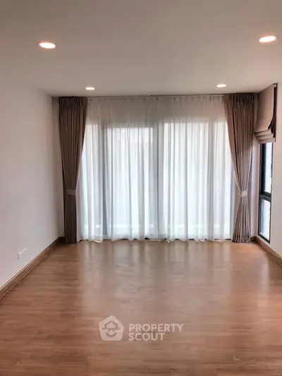 Spacious empty living room with wooden flooring and large windows with elegant curtains.