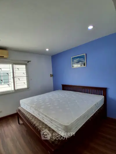Spacious bedroom with large window and blue accent wall, featuring a comfortable bed and wooden flooring.