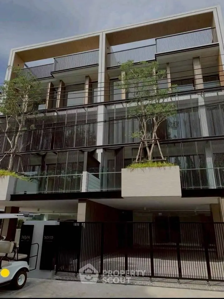Modern multi-story building with sleek design and balconies, featuring lush greenery and gated entrance.