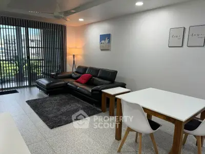Modern living room with sleek black leather sofa and stylish dining area, featuring large windows and natural light.