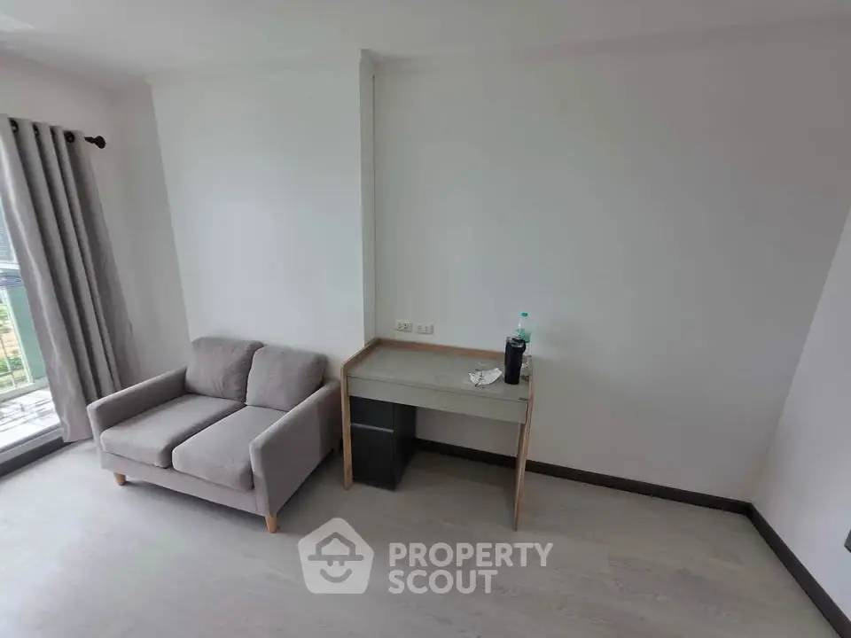 Minimalist living room with gray sofa and small desk, featuring large window with curtains.