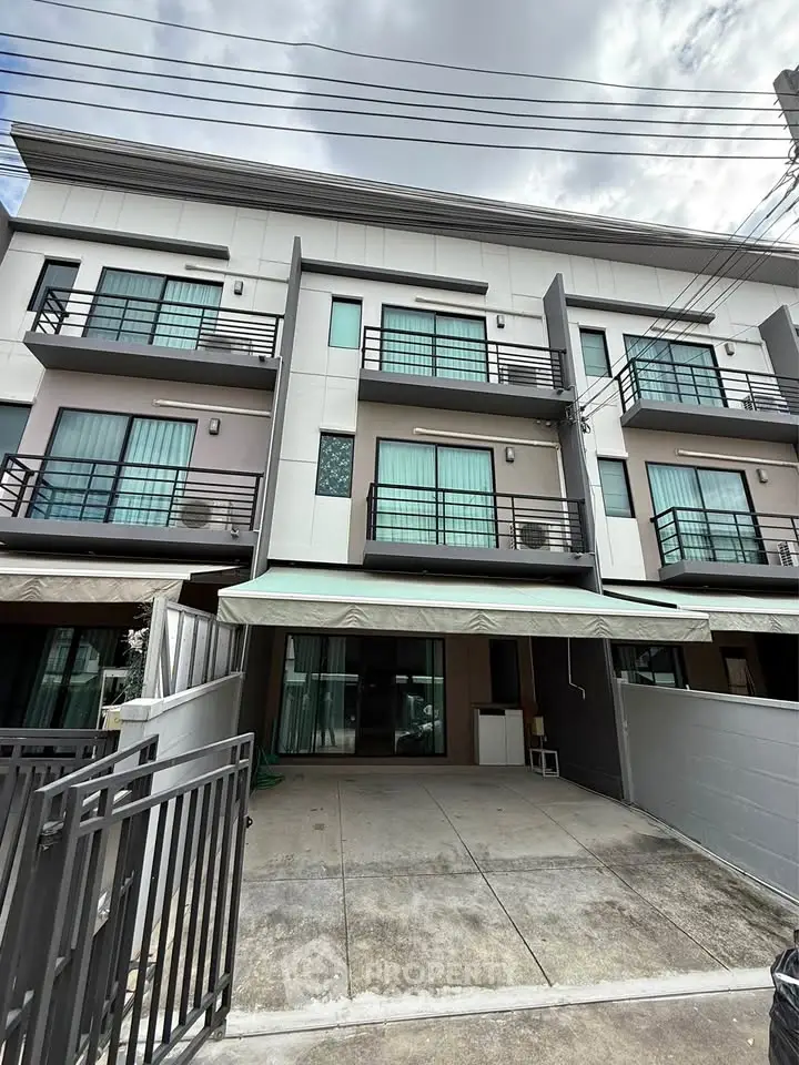 Modern three-story townhouse with balconies and gated parking area.