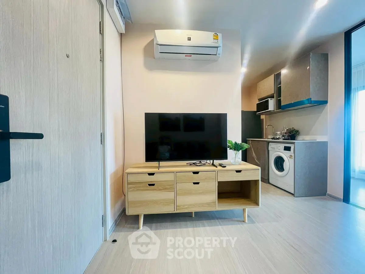 Modern living room with TV, air conditioning, and compact kitchen featuring washing machine.