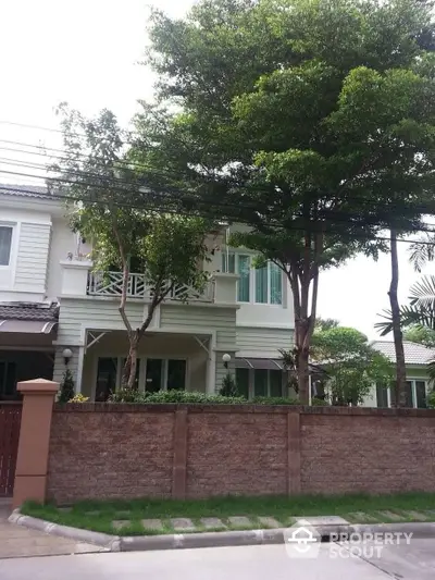 Charming two-story home with a lush green front yard, shaded by mature trees, featuring a spacious balcony and elegant white facade.