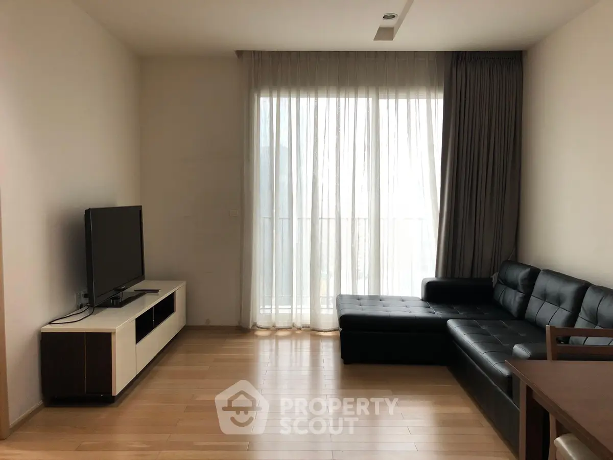 Spacious living room with modern black leather sofa and large TV, featuring natural light from floor-to-ceiling window.