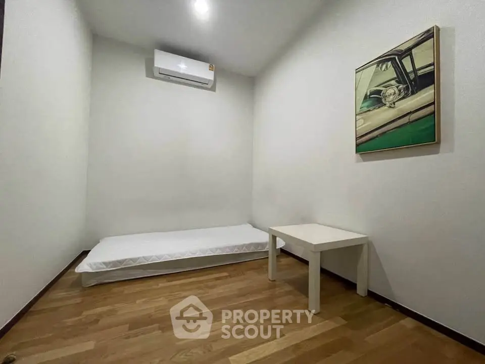 Minimalist bedroom with wooden flooring and air conditioning, featuring a single bed and small table.
