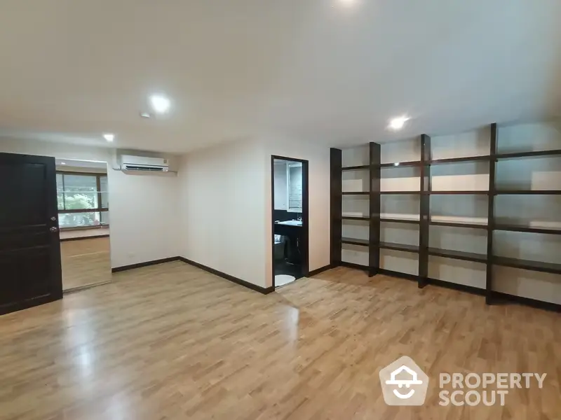 Spacious living room with polished wooden flooring, modern air conditioning unit, and built-in shelving, leading to a cozy kitchen area.