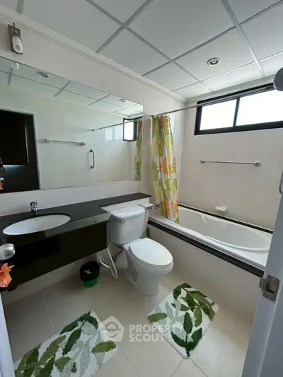 Modern bathroom with bathtub and large mirror, featuring tropical-themed decor and natural light.