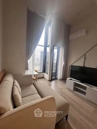 Modern living room with high ceilings and large windows offering city views, featuring a cozy sofa and sleek TV setup.