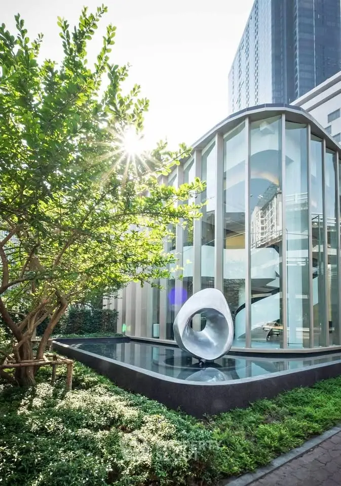 Modern architectural building with glass facade and artistic sculpture in lush garden setting.