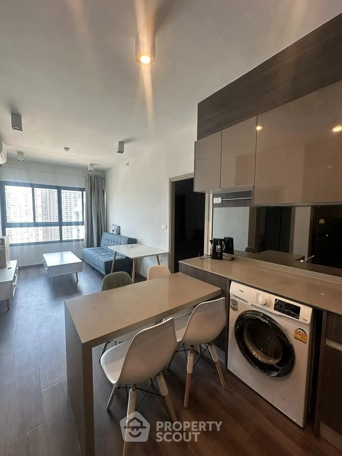 Modern living room with open kitchen and washing machine, featuring sleek furniture and ample natural light.