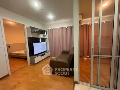 Modern living room with wooden flooring, cozy sofa, and large TV, adjacent to a bedroom with sliding glass doors.
