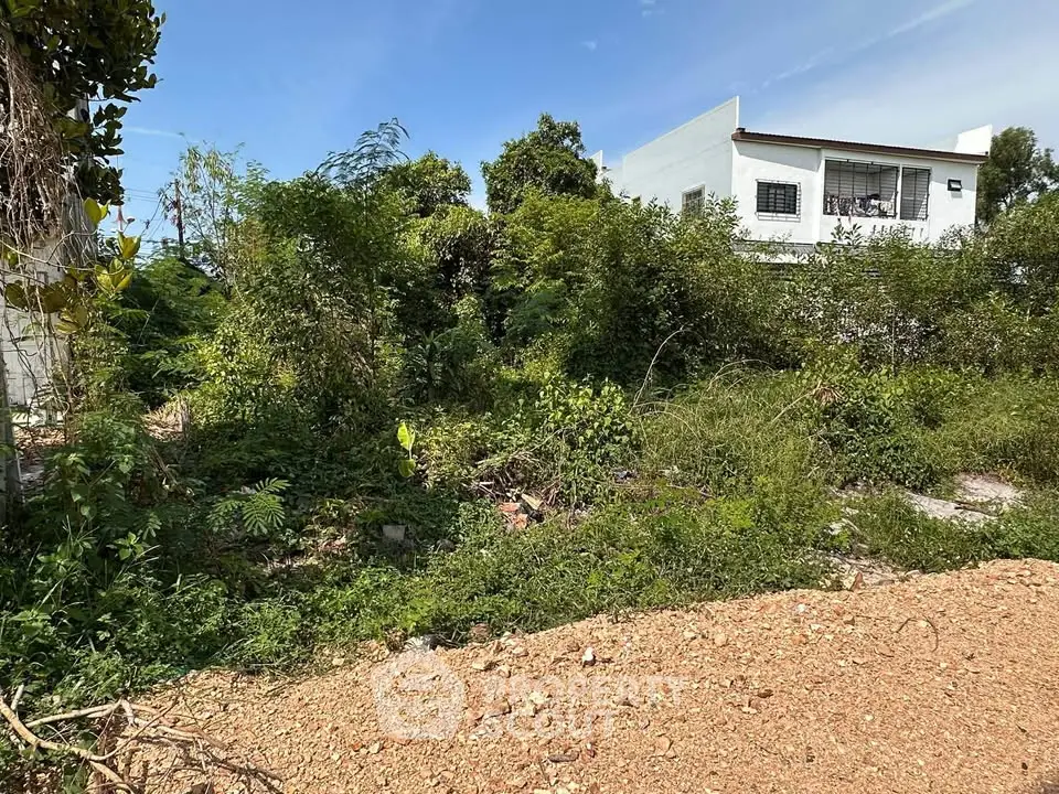 Vacant land plot with lush greenery and nearby residential building, ideal for development.