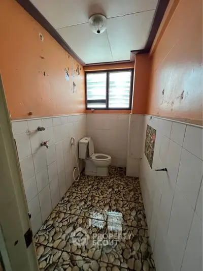 Compact bathroom with unique pebble stone flooring and orange walls, featuring a toilet and window for natural light.
