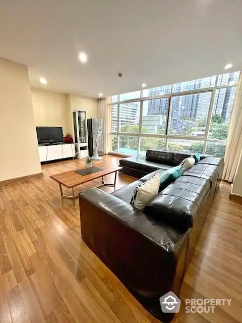 Spacious living room with large windows and city view, featuring modern leather sofa and wooden flooring.