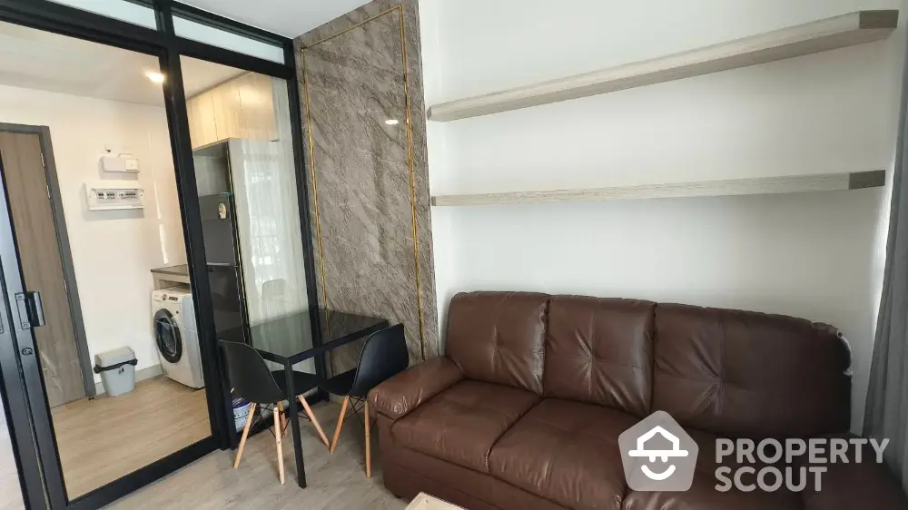 Modern living room with brown leather sofa and glass dining table adjacent to kitchen area.