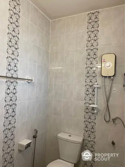 Compact bathroom with modern electric shower unit and neutral tile design, featuring a clean white toilet and convenient safety grab bars.