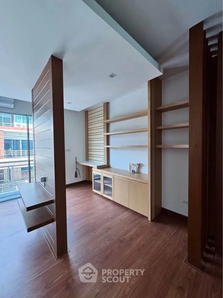 Modern living room with wooden flooring and built-in shelves, featuring large windows for natural light.