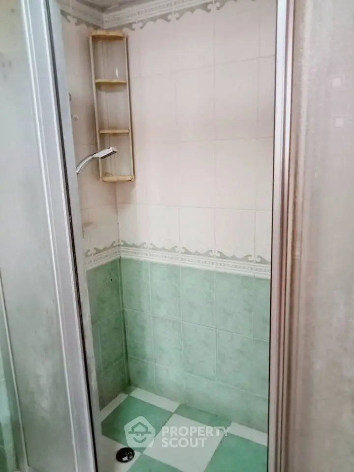 Compact bathroom shower area with green and white tiles, featuring a glass door and corner shelving.