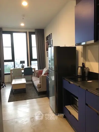 Modern open-plan living room with kitchen, featuring sleek blue cabinets and large windows.