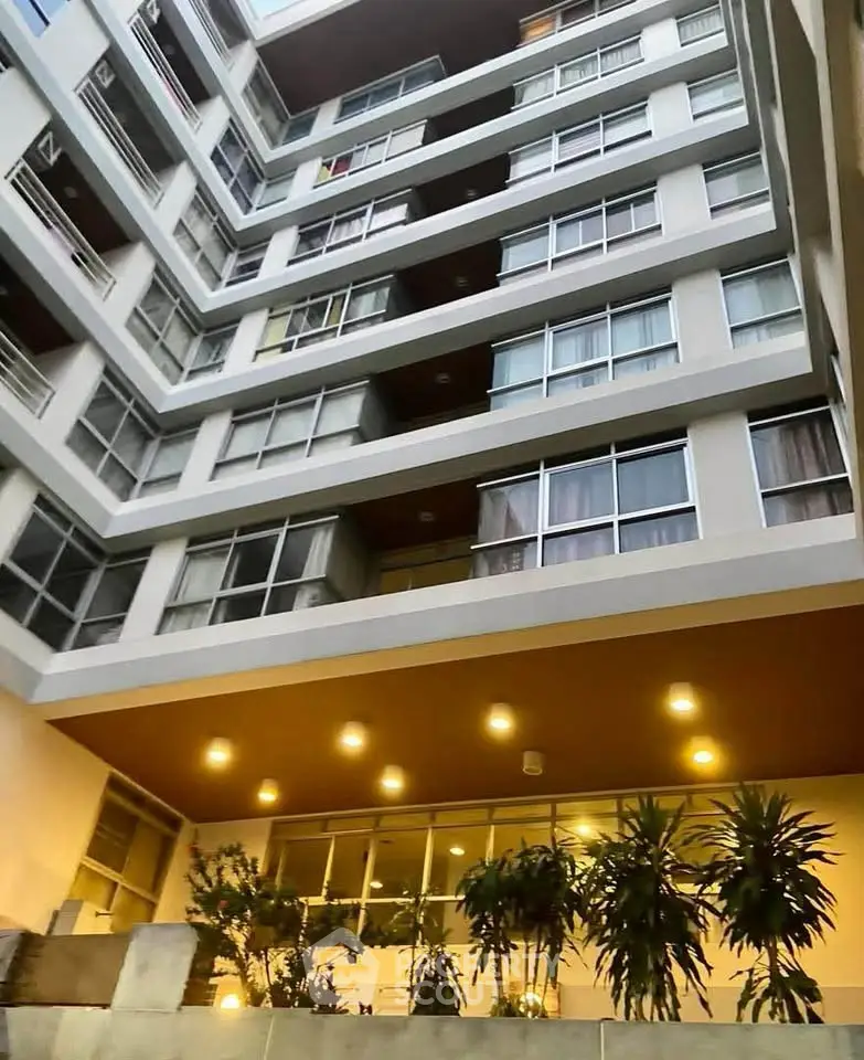 Modern apartment building with large windows and balcony views, featuring a well-lit entrance and lush greenery.