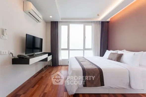 Modern bedroom with wooden flooring, large window, and sleek TV setup in a stylish apartment.
