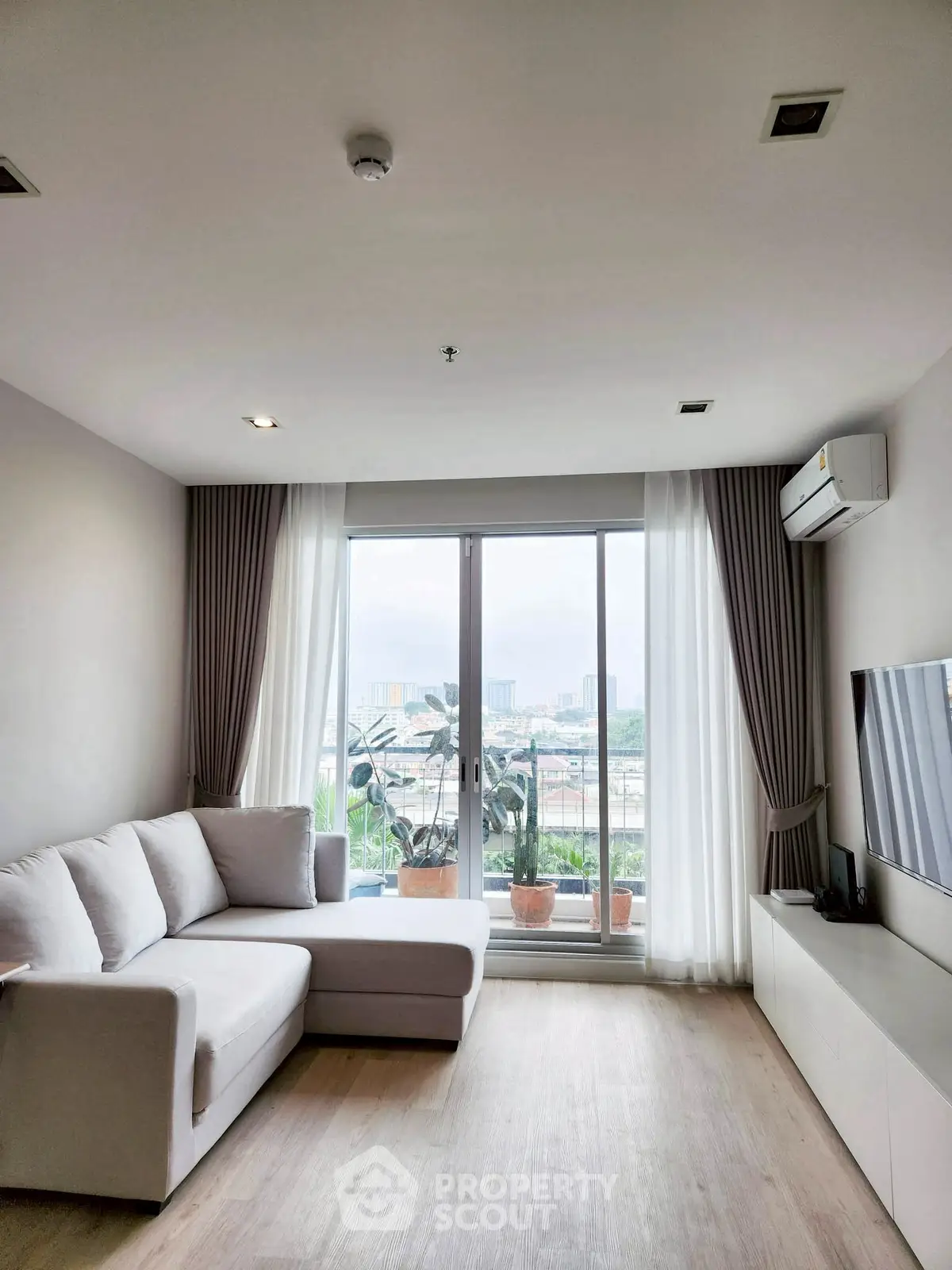 Modern living room with large window and city view, featuring a cozy white sofa and sleek TV setup.