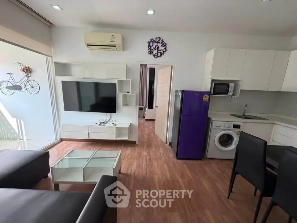 Modern living room with sleek furniture, large TV, and kitchen appliances in open layout.