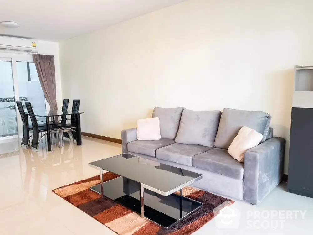 Spacious living room with glossy tiled floors, modern grey sofa, elegant glass coffee table, and a sleek dining set by the window, exuding contemporary charm.