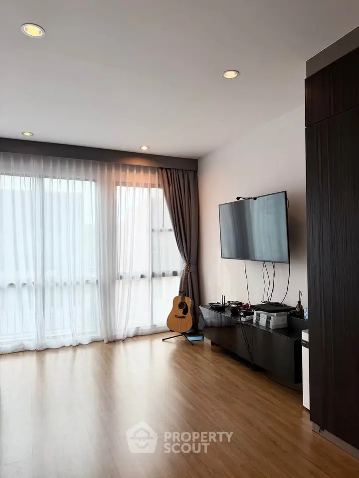 Spacious living room with wooden flooring and modern decor, featuring a wall-mounted TV and large windows.