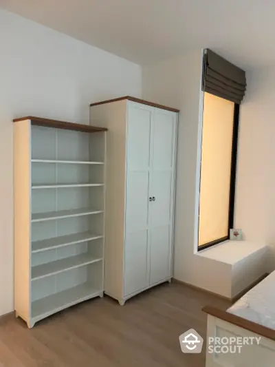 Bright and cozy bedroom with ample storage, featuring a large wardrobe and a bookshelf, complemented by warm wooden flooring and a large window for natural light.