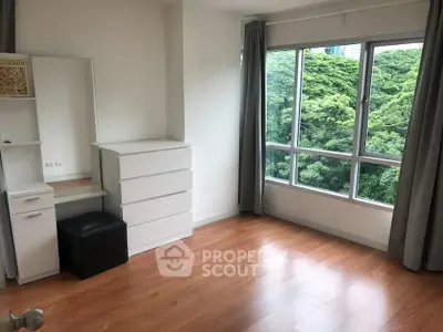 Bright bedroom with large windows and wooden flooring, offering a serene view of lush greenery.