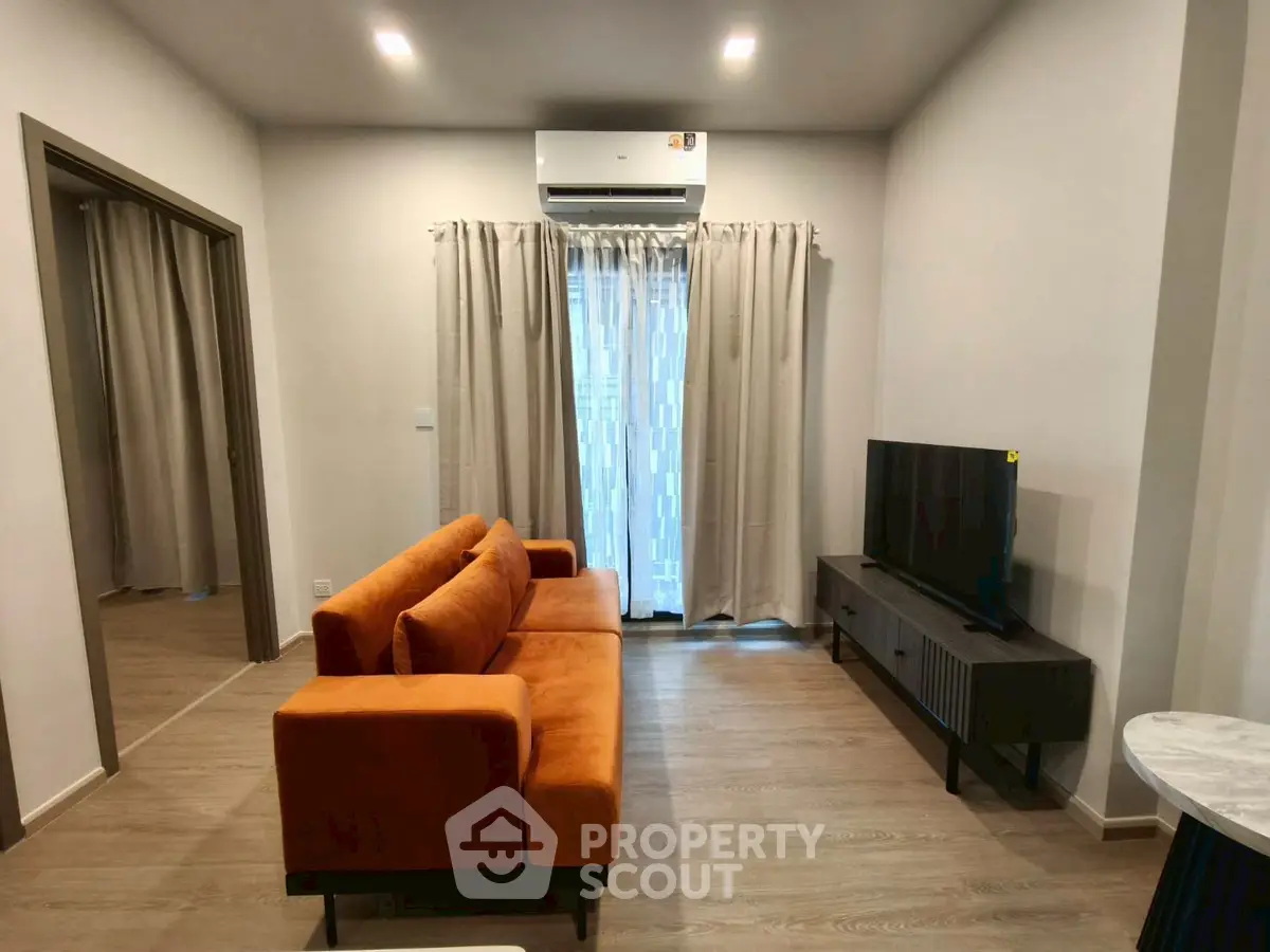 Modern living room with orange sofa, TV, and air conditioning, featuring wooden flooring and neutral curtains.