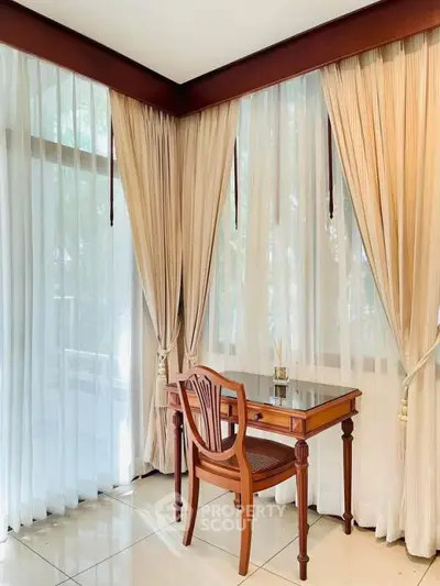 Elegant study nook with wooden desk and chair by large windows with sheer curtains.