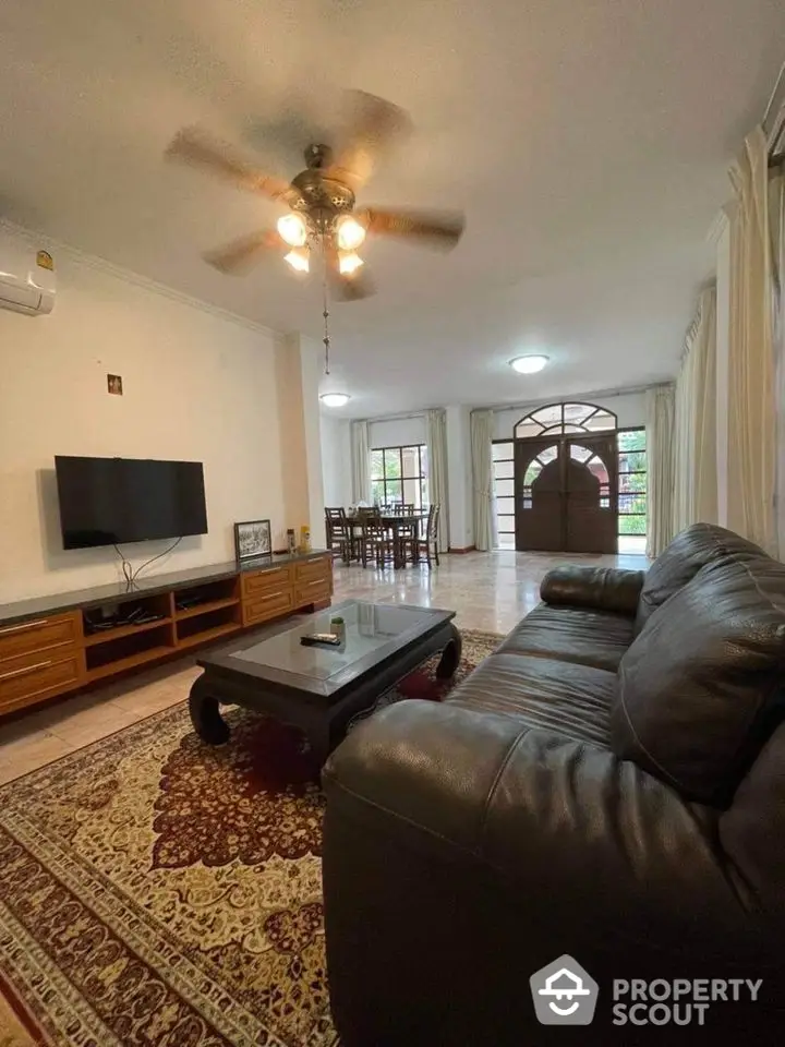 Spacious living room with polished tile flooring, plush leather sofa, and a classic ceiling fan, leading to a bright dining area with archway entrance.