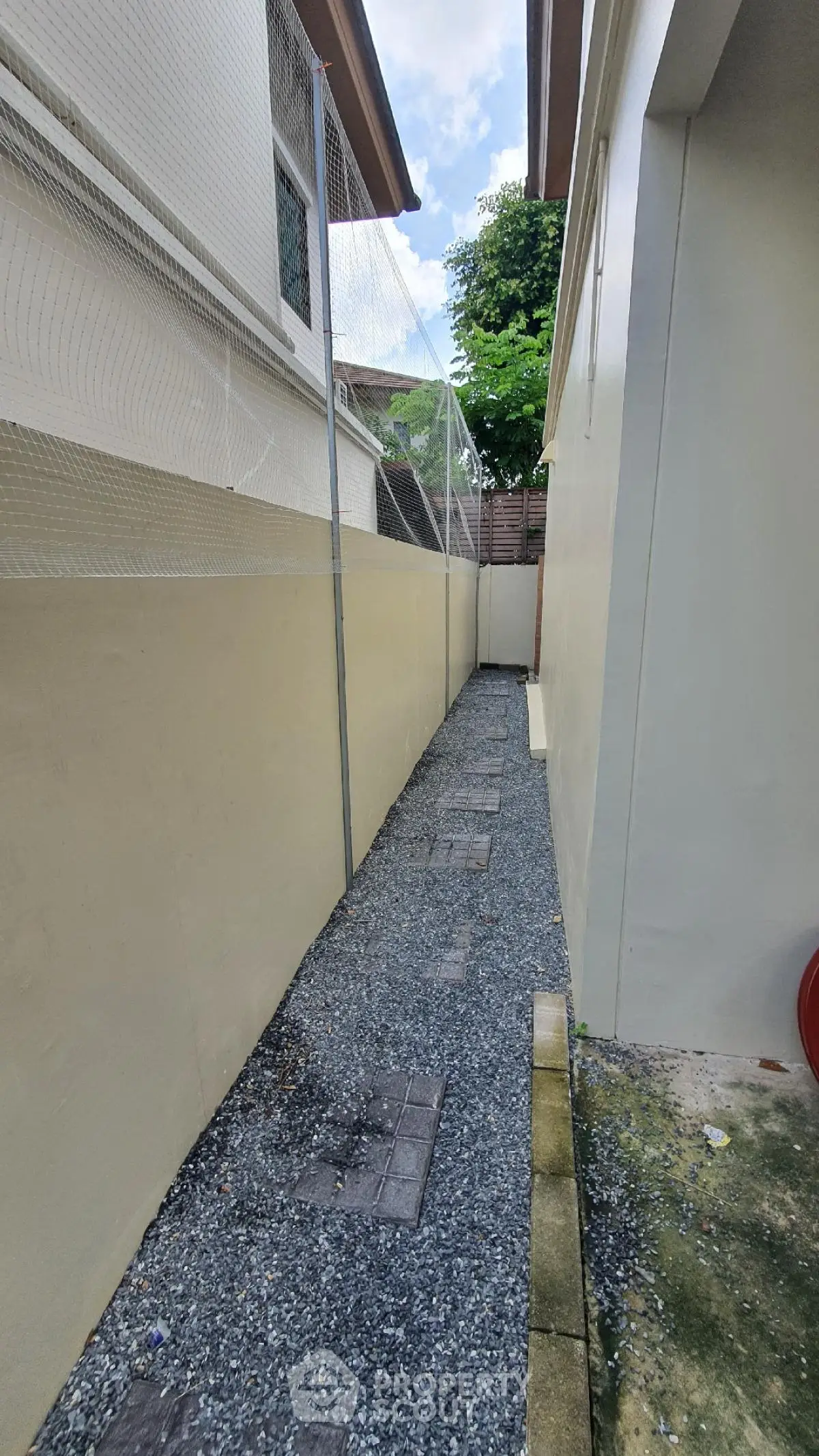 Narrow outdoor walkway with gravel path between two buildings, featuring a netted fence and clear sky above.