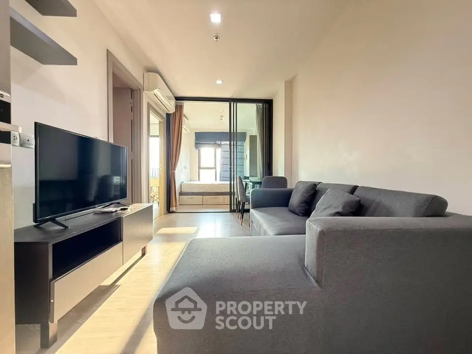 Modern living room with sleek gray sofa and flat-screen TV, leading to a cozy bedroom through glass doors.